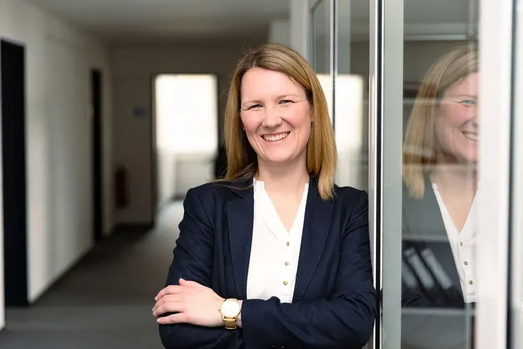 Jennifer Guenther lächelt im Business-Outfit und steht an einer Glaswand im Büro