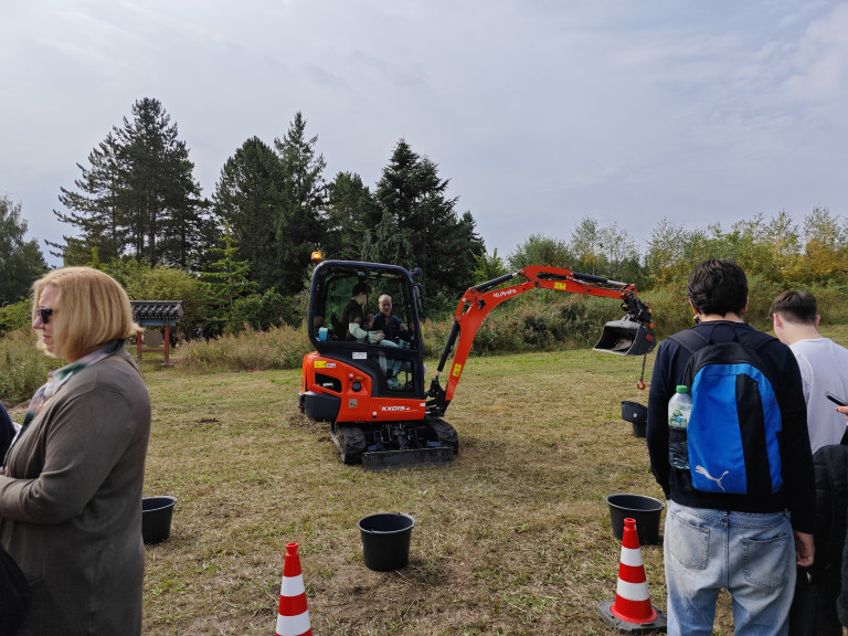 Bau-Event mit Minibagger und Personen auf einer Wiese Bau-Event mit Minibagger auf einer Wiese, Personen bedienen den Bagger, Baukegel und Eimer im Vordergrund