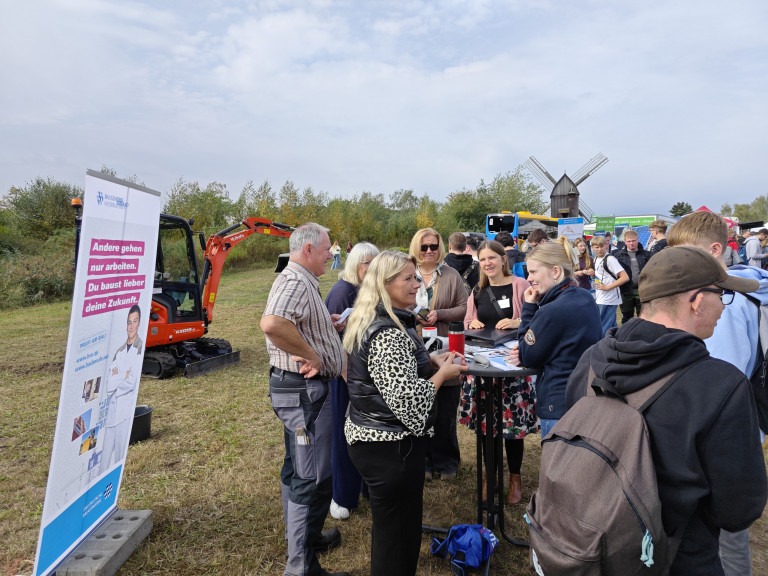 Informationsstand Bauwesen auf Messe im Freien mit Bagger und Windmühle Informationsstand zum Bauwesen auf einer Messe im Freien mit Menschen im Gespräch, Bagger und Windmühle im Hintergrund