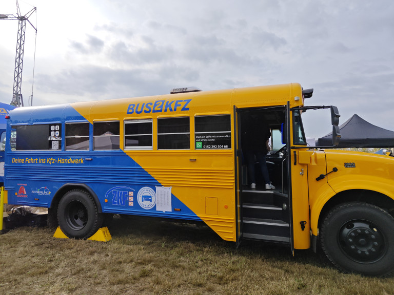 BUS2KFZ Bus präsentiert Kfz-Handwerk auf Event Gelber und blauer Bus mit der Aufschrift BUS2KFZ, präsentiert das Kfz-Handwerk auf einer Veranstaltung.