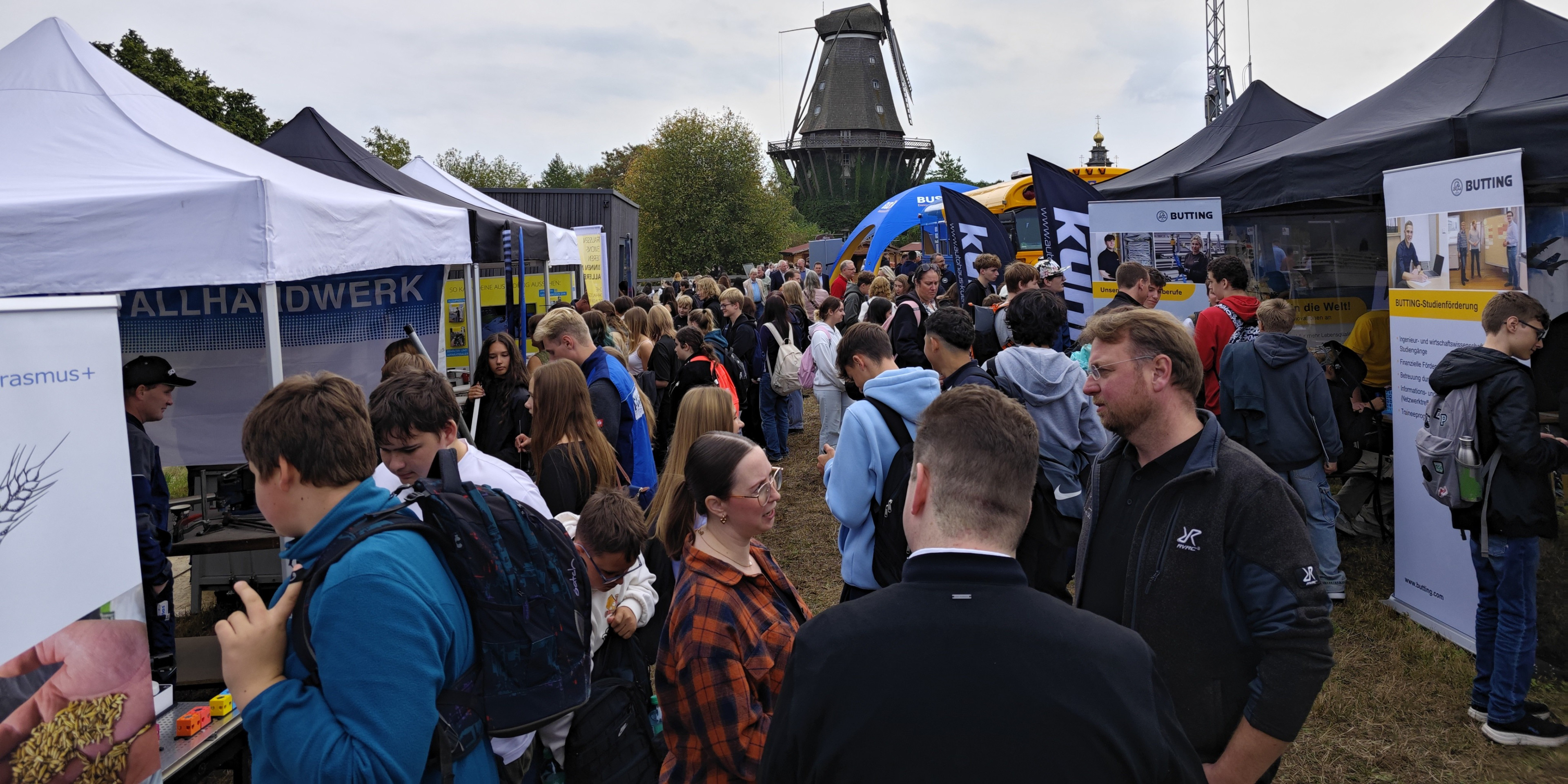 Viele Menschen besuchen eine Messe mit Informationsständen im Freien, im Hintergrund ist eine Windmühle zu sehen.