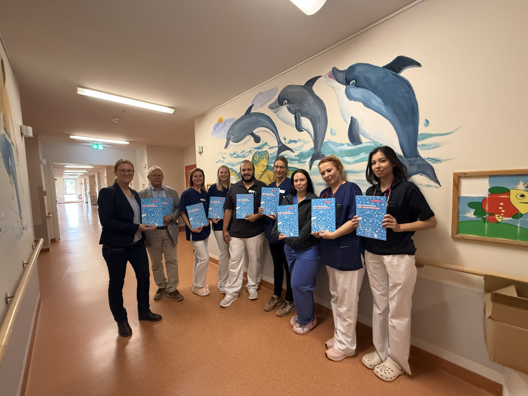 Team des Helios Klinikums Gifhorn mit Informationsmaterial vor Delfin-Wandbild Teamfoto im Helios Klinikum Gifhorn vor Wandbild mit Delfinen, Mitarbeitende halten Informationsmappen in den Händen.