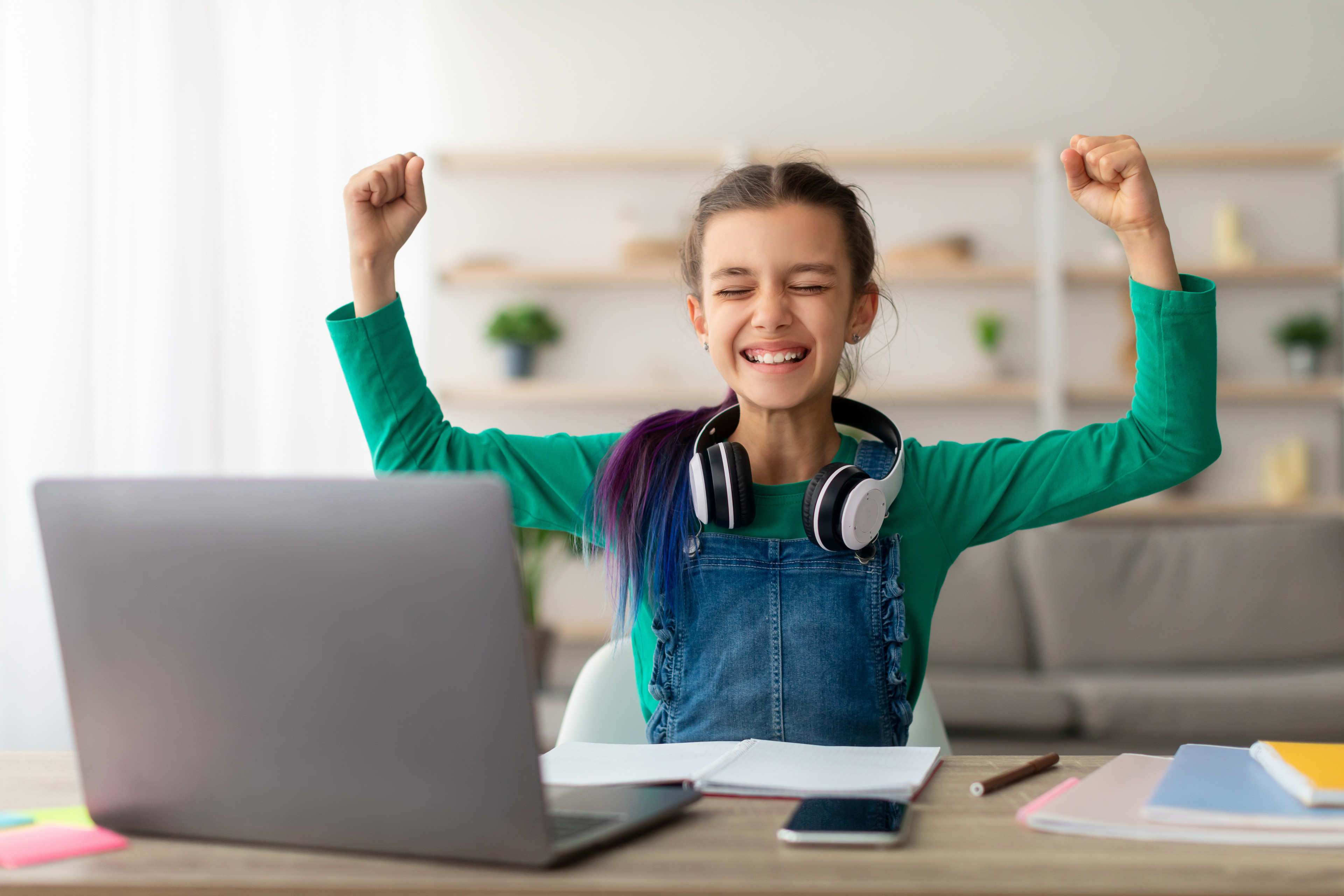 Mädchen mit Kopfhörern lernt erfolgreich am Laptop und hebt die Arme zum Jubeln