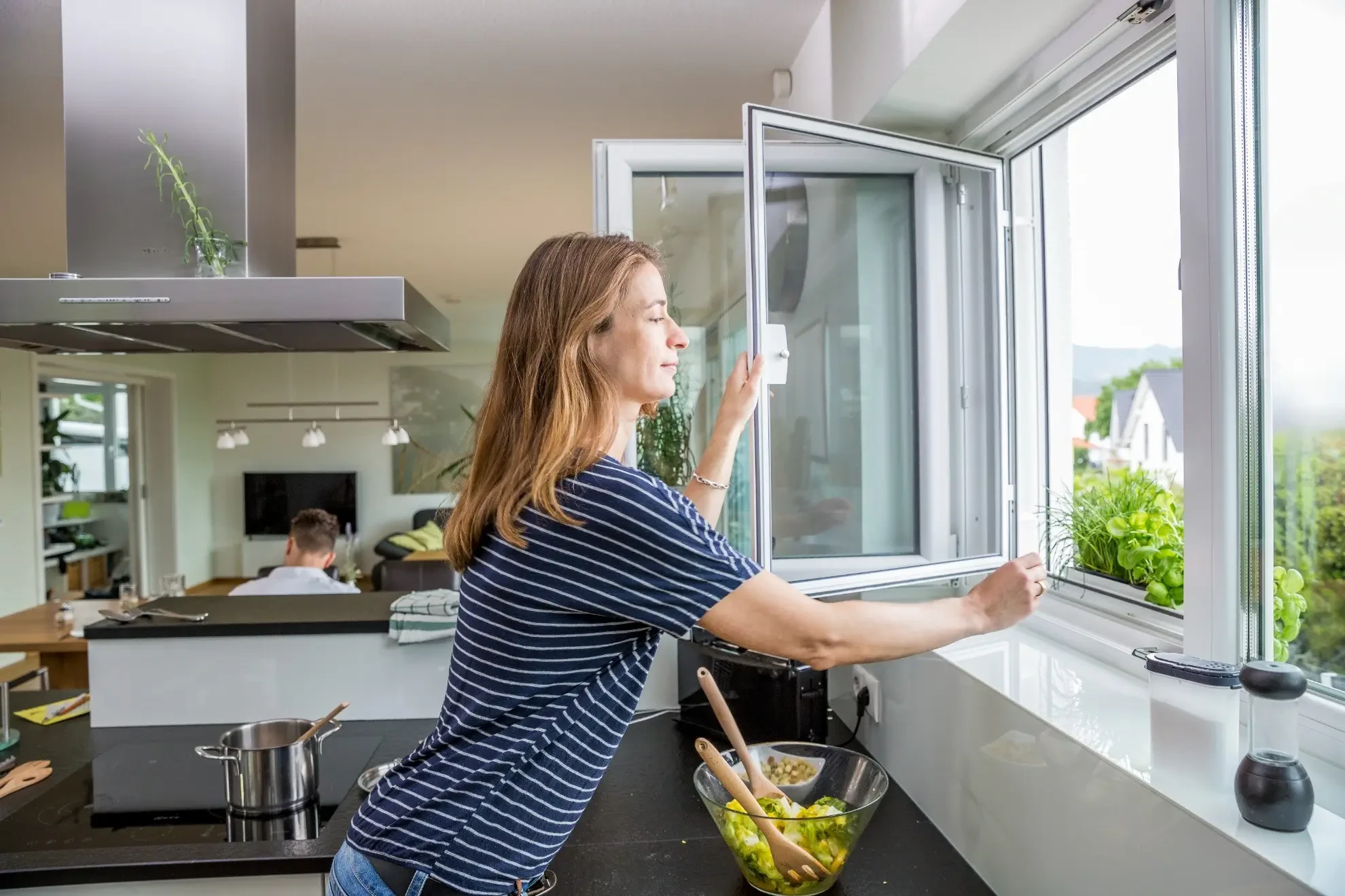 Frau öffnet ein Fenster in einer modernen Küche mit Salatschüssel auf der Arbeitsplatte