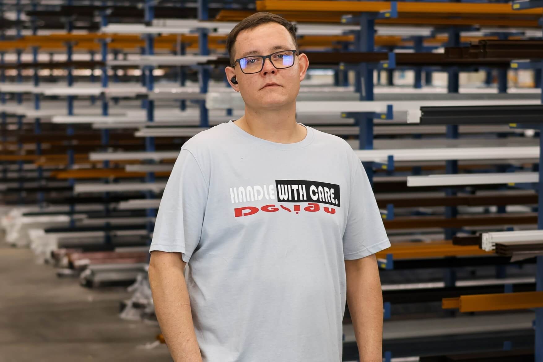 Mann im Lager in weißem T-Shirt mit der Aufschrift 'Handle with care' und Brille.