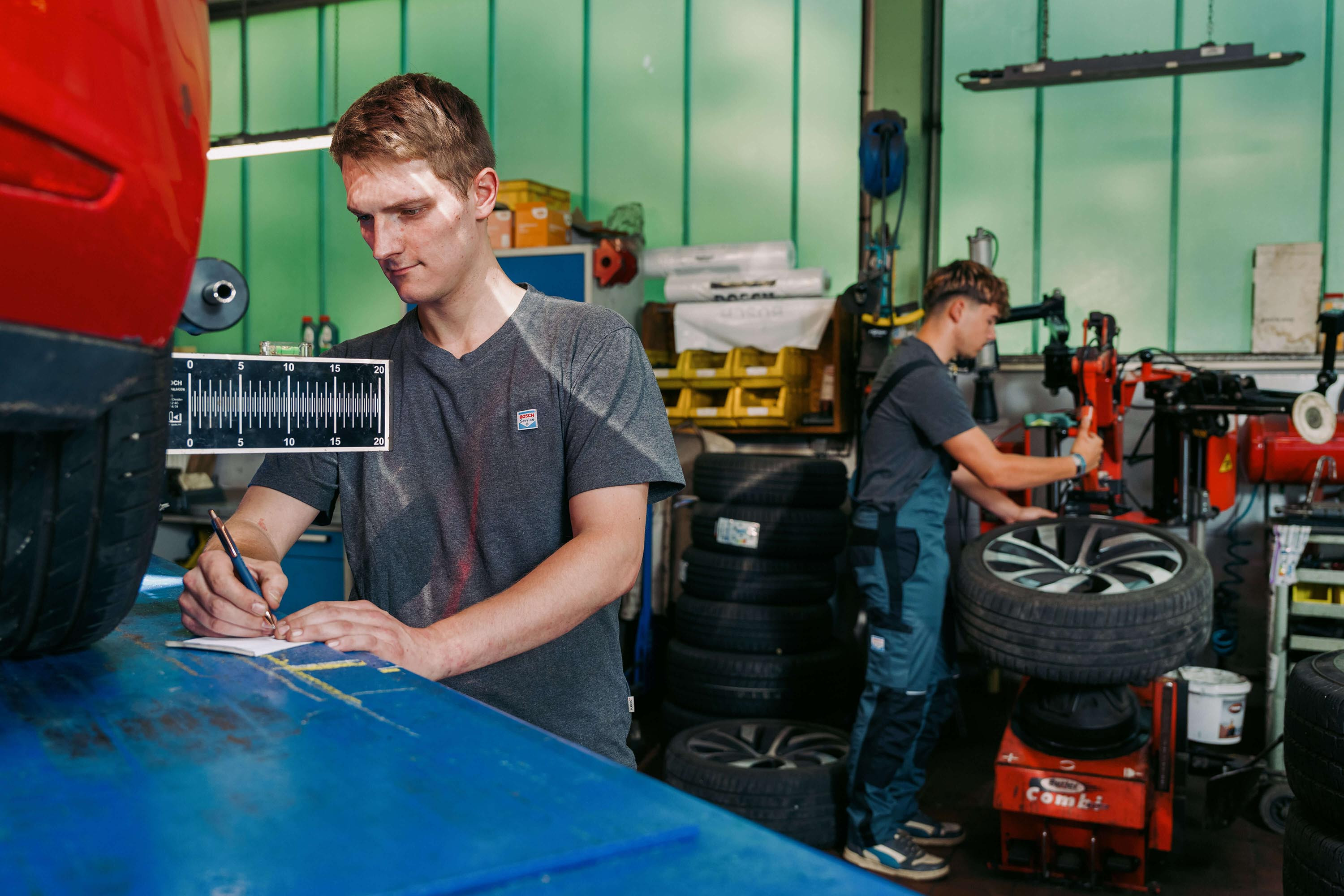 Zwei Kfz-Mechaniker bei der Arbeit in einer Autowerkstatt, einer schreibt am Tresen, der andere montiert ein Rad.