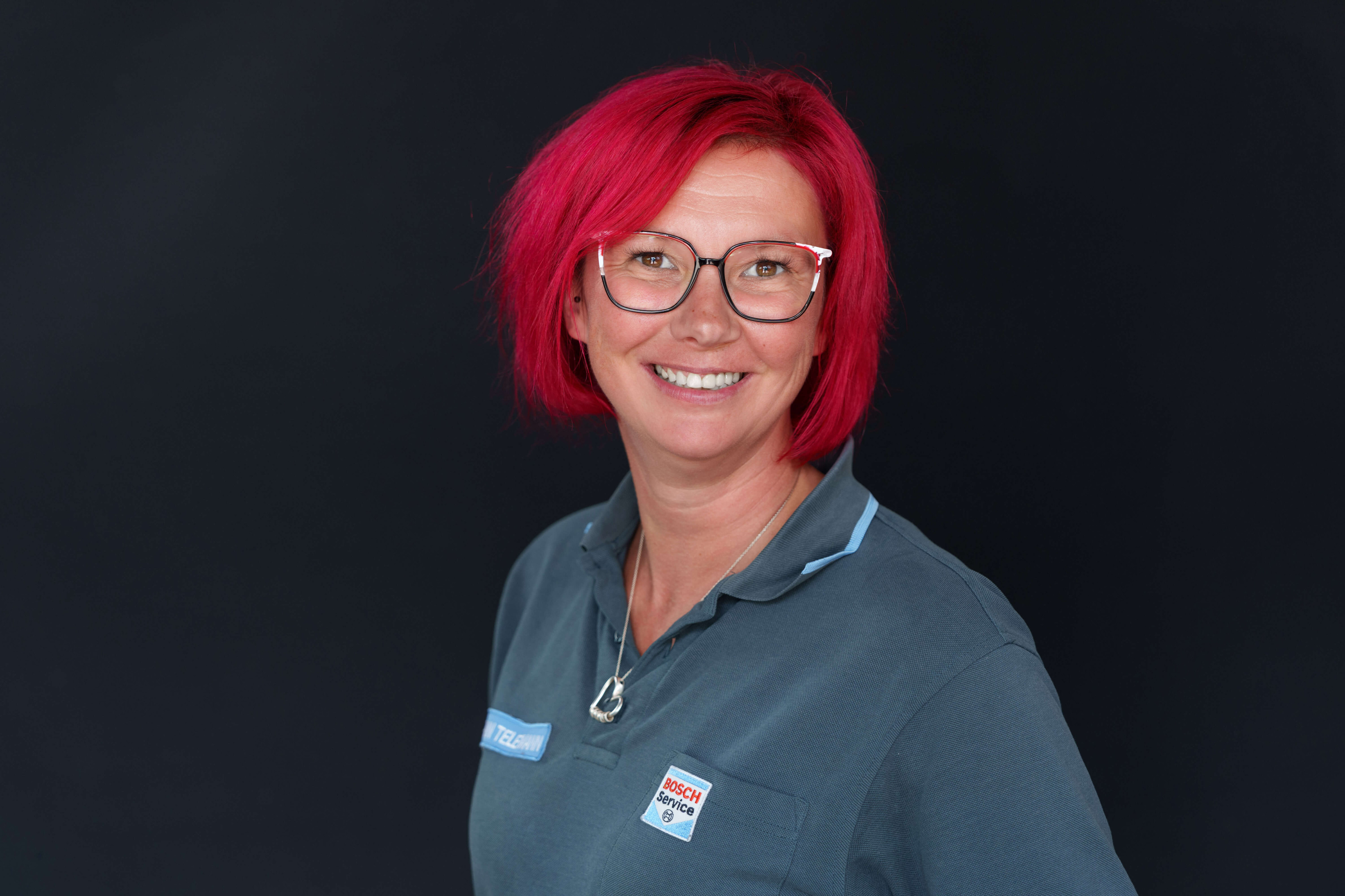 Mitarbeiterin mit roten Haaren und Brille im Bosch Service Outfit vor dunklem Hintergrund