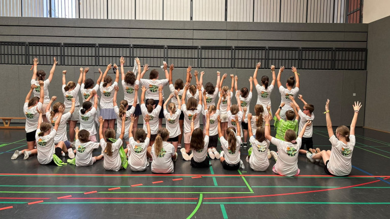 Gruppenfoto vom Sommercamp in der Sporthalle – Kinder und Jugendliche mit erhobenen Armen Kinder und Jugendliche beim Sommercamp in einer Sporthalle, Gruppenfoto von hinten mit erhobenen Armen