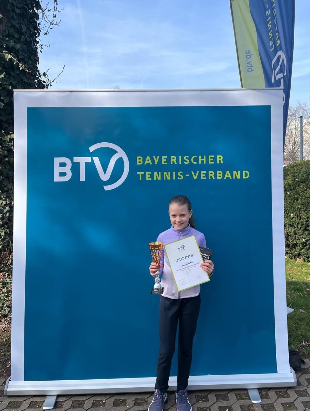 Vanessa bei der Bayerischen Meisterschaft 2026 mit Pokal und Urkunde vor dem Banner des Bayerischen Tennis-Verbands