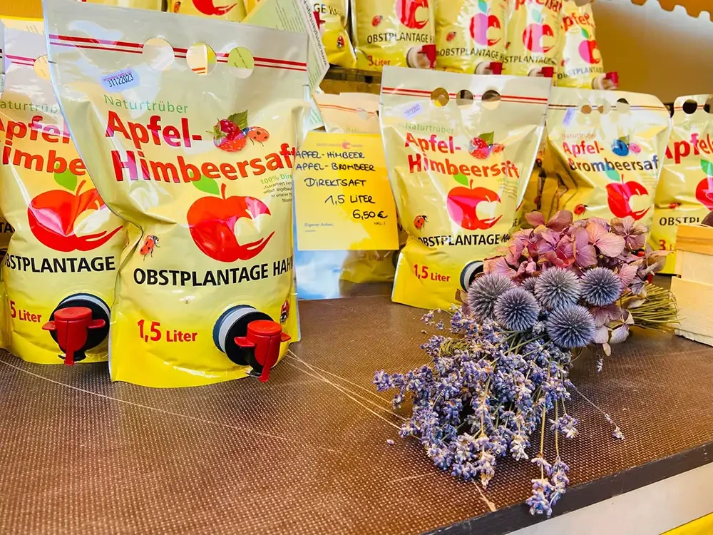 Apfel-Himbeersaft Direktsaft von Obstplantage Hahn – regionale Verarbeitungsprodukte im Verkauf Direktsaft Apfel-Himbeersaft von Obstplantage Hahn in 1,5 Liter Verpackungen auf einem Verkaufstisch mit Trockenblumen