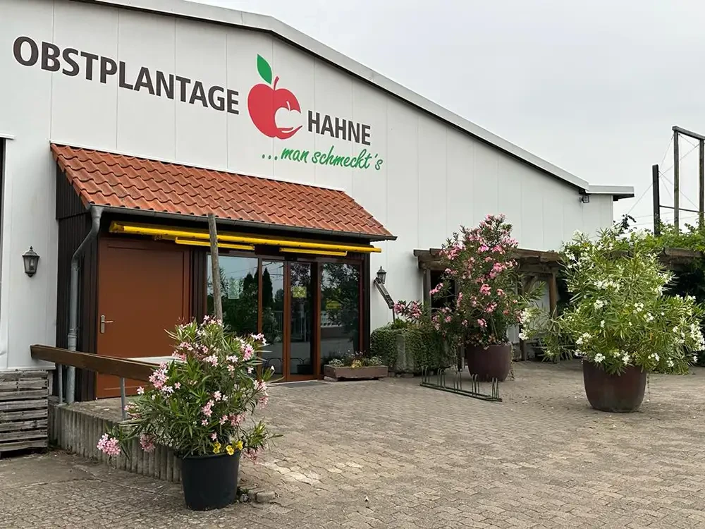 Hofladen der Obstplantage Hahne – Regional einkaufen und genießen Eingangsbereich des Hofladens der Obstplantage Hahne mit blühenden Pflanzen und Firmenschild