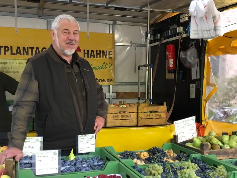 Frischer Obststand auf dem Wochenmarkt – regionale Produkte bei Hahne Marktstand mit frischem Obst auf dem Wochenmarkt, Verkäufer hinter dem Stand, Schild mit 'Hahne' im Hintergrund
