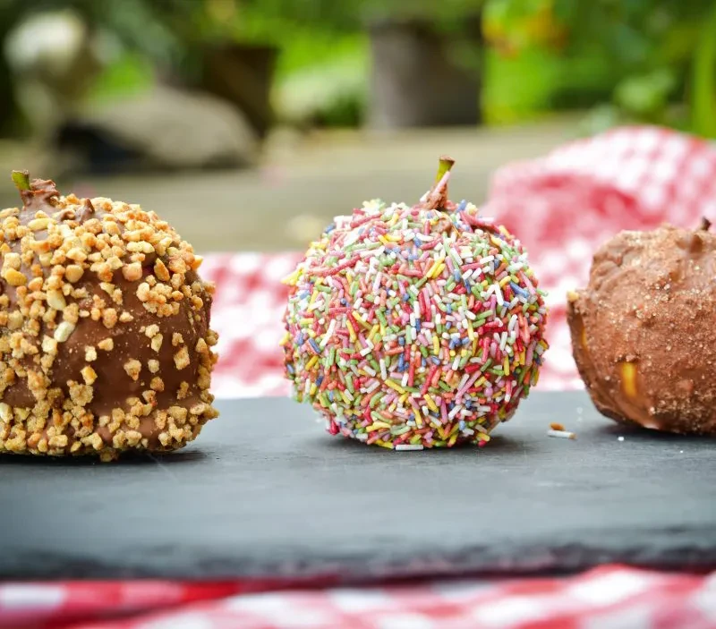 Apfel-Zauberspieße mit Schokolade, Nüssen und Streuseln als kreative Snack-Idee Bunte Apfel-Zauberspieße mit Schokolade, Nüssen und Zuckerstreuseln auf einer Schieferplatte