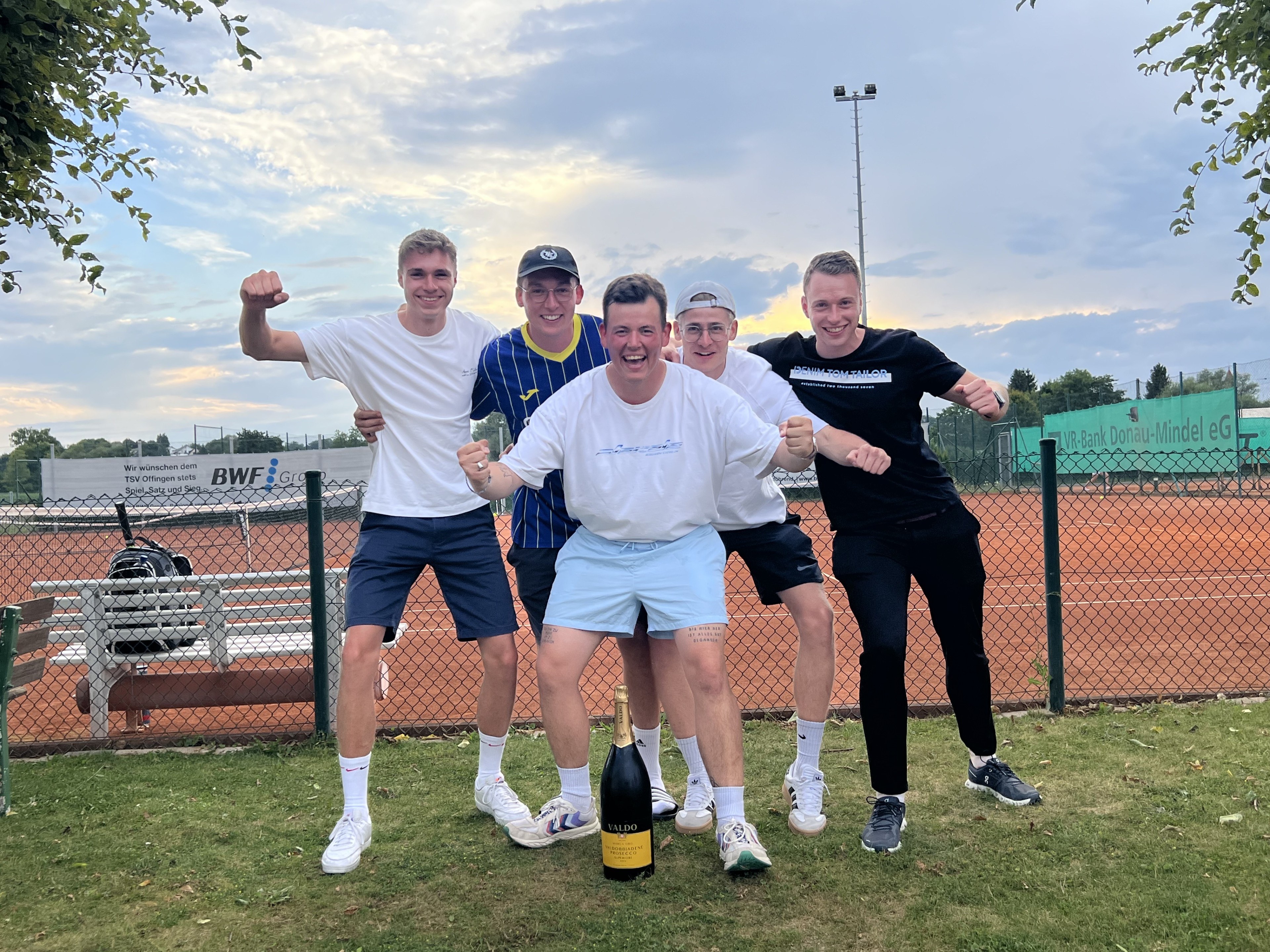 Gruppe von jungen Männern auf einem Tennisplatz mit einer Champagnerflasche