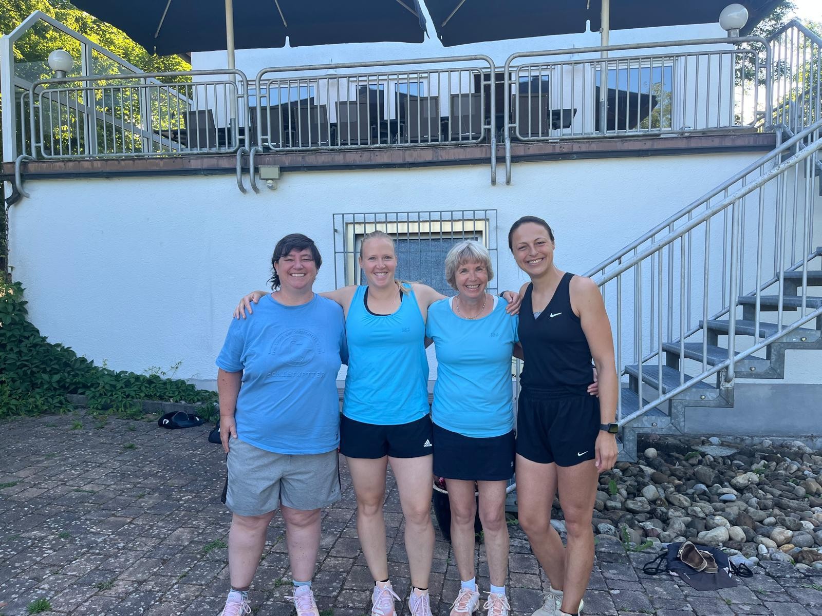 Vier Frauen in Sportkleidung stehen gemeinsam vor einem Vereinsgebäude und lächeln in die Kamera. Sommerliche Szene, Gruppenfoto nach dem Sport.