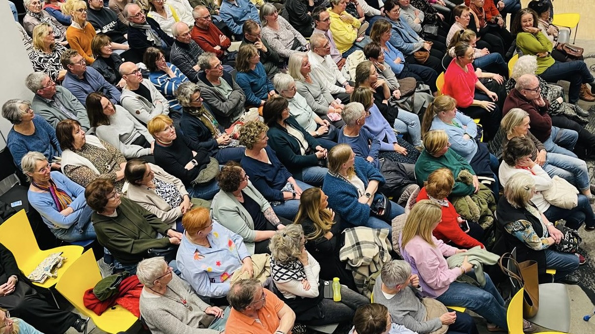 Viele Menschen sitzen dicht beieinander bei einer Veranstaltung oder einem Vortrag in einem Saal.
