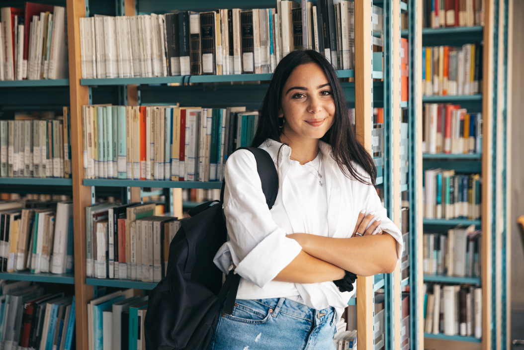 Studentin mit Rucksack steht vor Bücherregal in einer Bibliothek