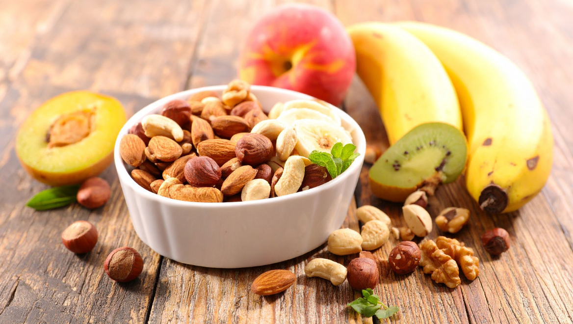 Schale mit Nüssen und Trockenfrüchten, daneben frische Banane, Kiwi und Pfirsich auf Holztisch