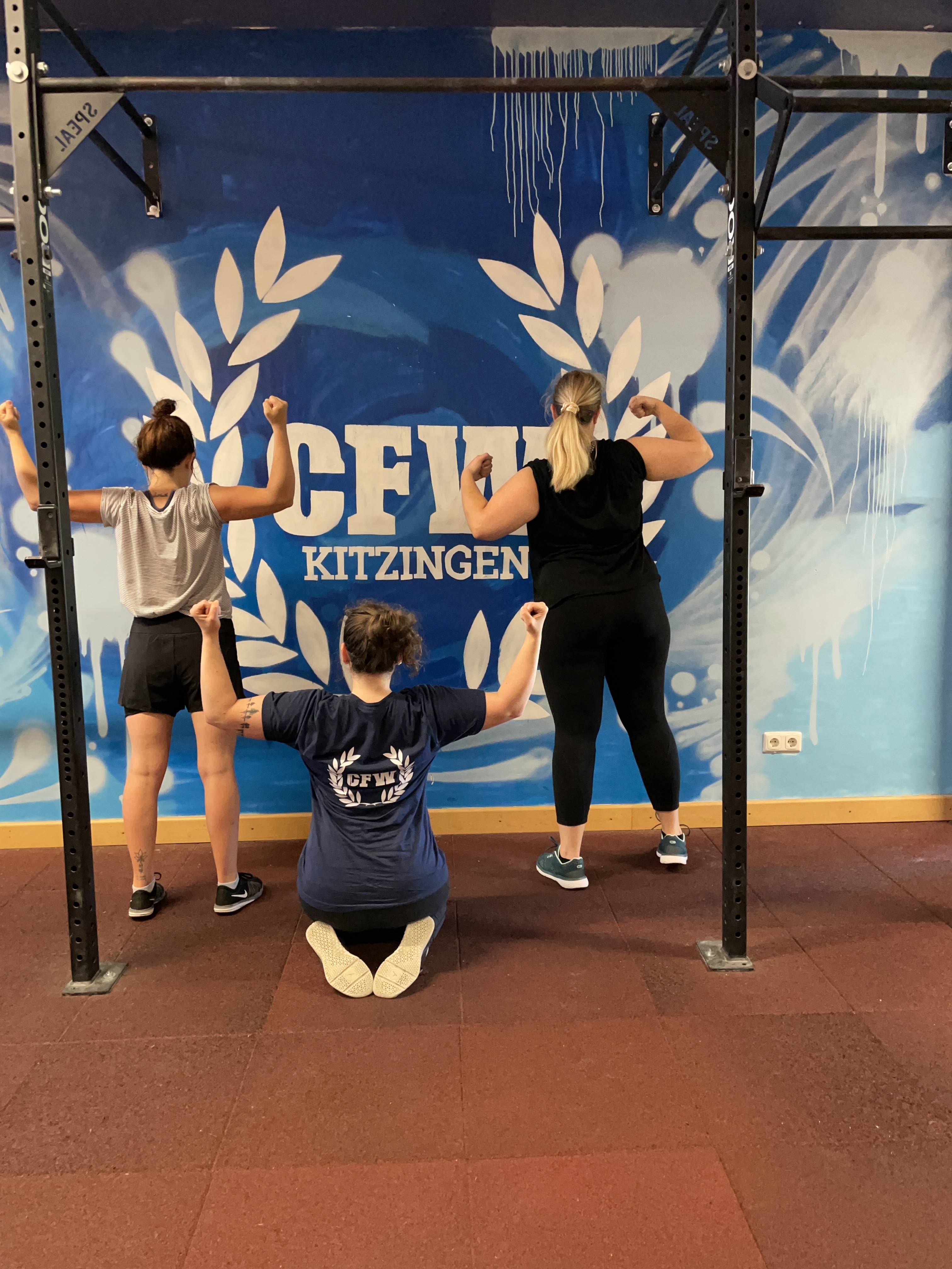 Drei Personen zeigen Muskeln vor einer CrossFit-Wand im Fitnessstudio.