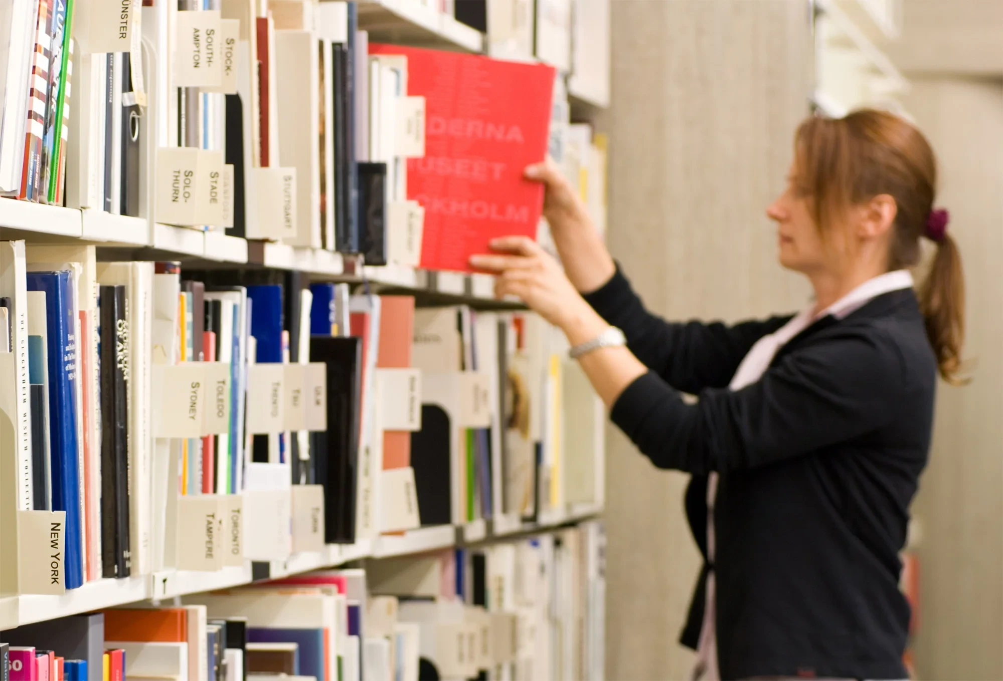 Frau stellt ein Buch ins Regal einer Bibliothek – beitrag-bibliothek