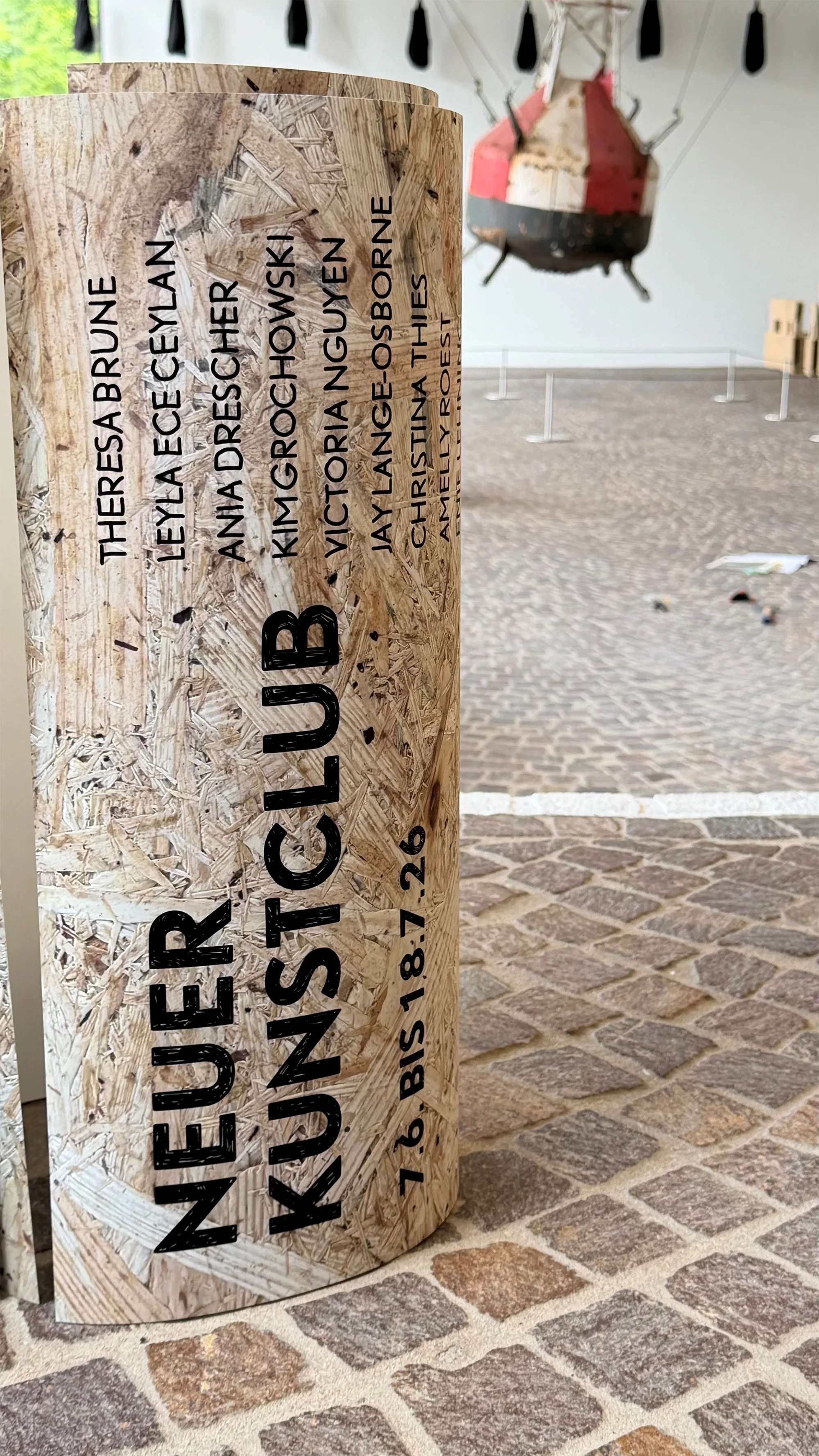 Plakat in der Lounge mit Künstlernamen und dem Titel 'Neuer Kunstclub', aufgenommen auf einer Pflasterfläche
