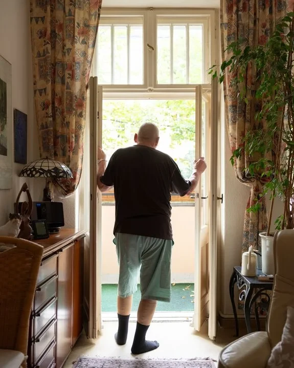 Älterer Mann steht in seinem Zuhause am offenen Balkon, Innenaufnahme mit gemütlicher Einrichtung