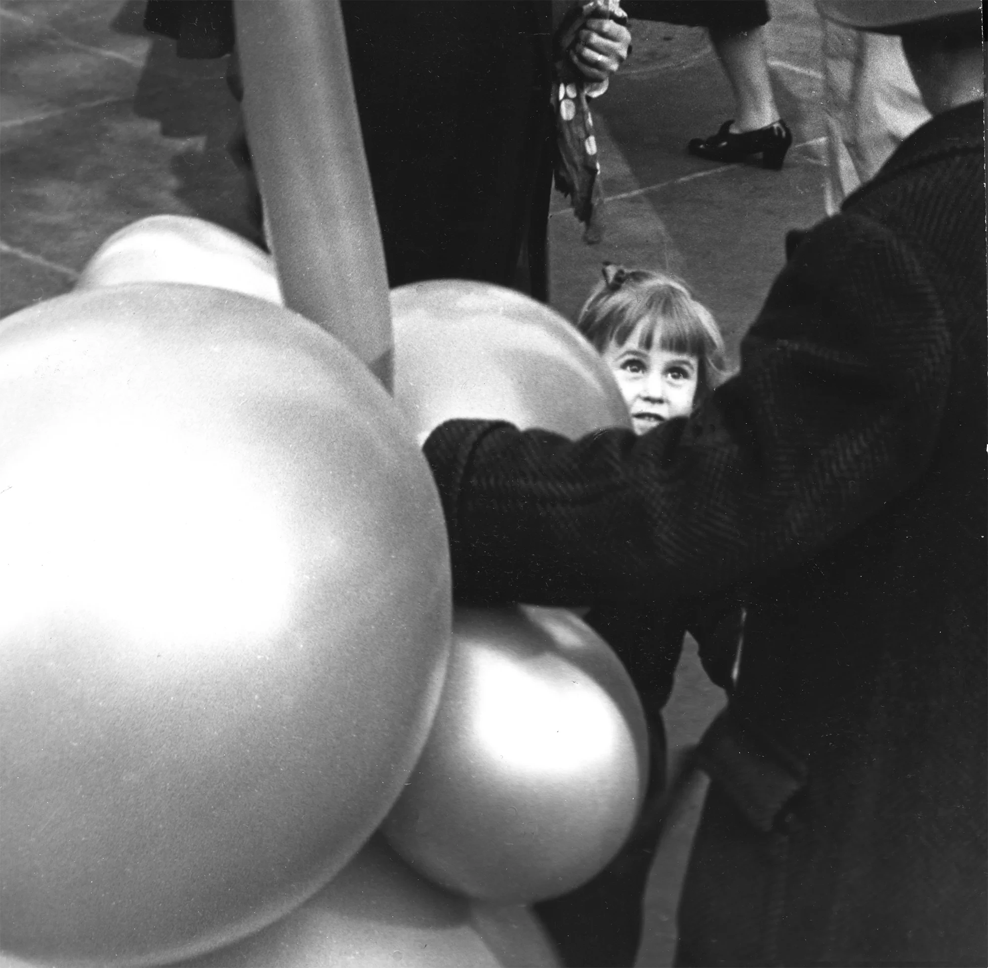 Schwarzweiß-Fotografie von Fred Stein: Ein kleines Mädchen blickt fasziniert auf große Luftballons in einer Straßenszene.