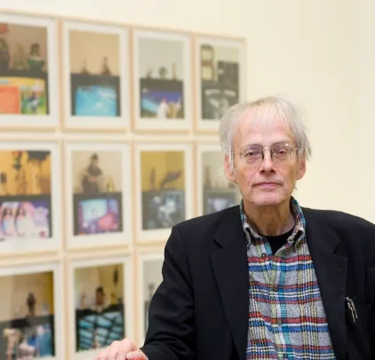 Älterer Mann mit Brille steht vor einer Wand mit gerahmten Fotografien, aufgenommen am 09. September 2026 um 09:38:27