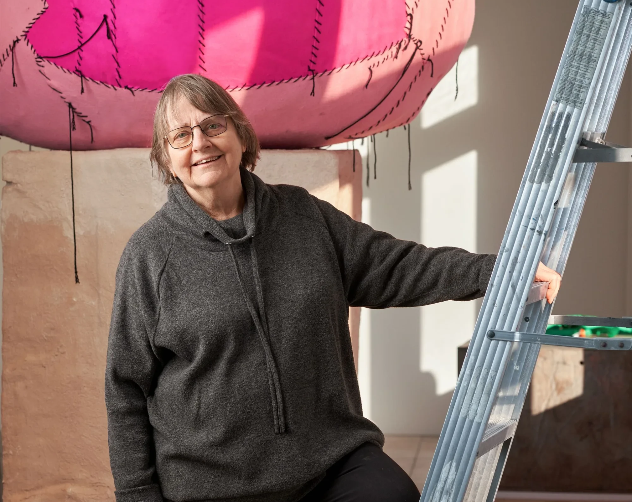Ältere Frau mit Brille steht neben einer Leiter vor einer Kunstinstallation mit pinkem Stoff am 09. September 2026