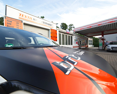 Auto vor Textilwaschstraße und Tankstelle, Nahaufnahme mit auffälligem Branding auf der Motorhaube