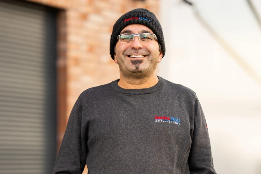 Hanifi Maurer in Arbeitskleidung vor einer Backsteinwand Hanifi Maurer mit Mütze und Brille vor einer Backsteinwand