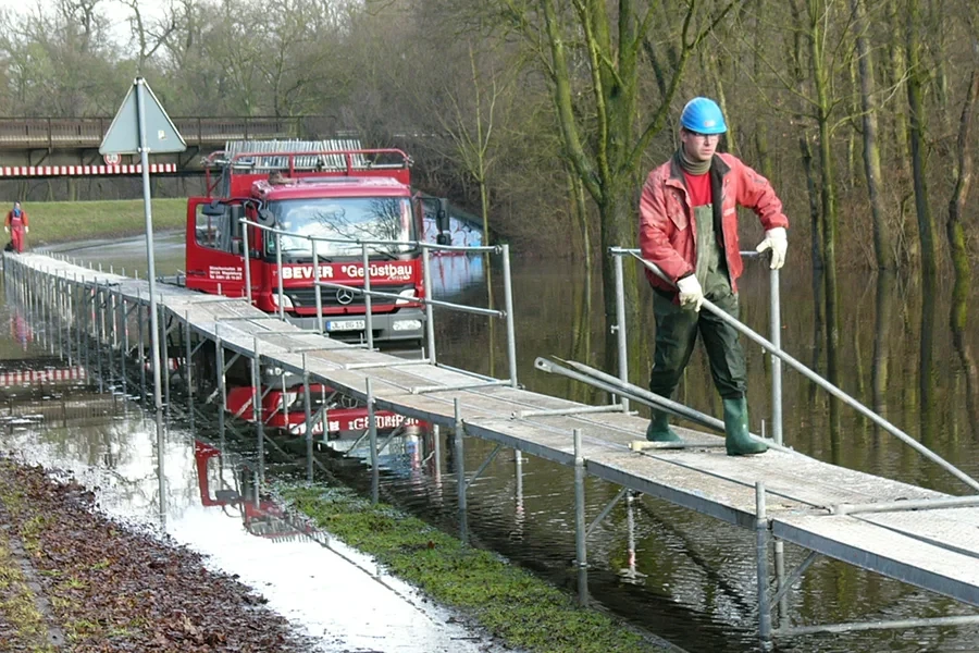 Karriere im Gerüstbau – Arbeiten bei BEVER Gerüstbau auf Baustelle im Freien Arbeiter mit Schutzhelm auf Gerüstbau-Brücke vor rotem BEVER Gerüstbau LKW im Freien