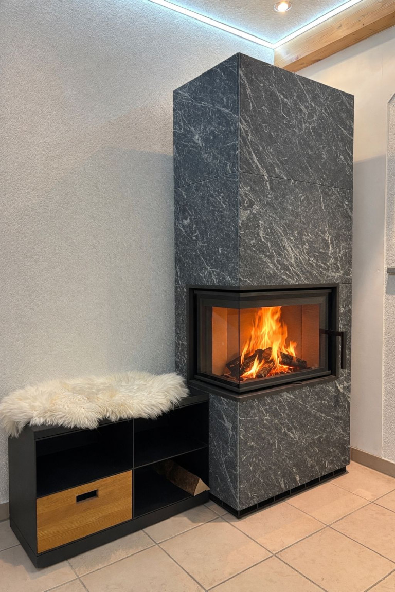 Moderner Kamin mit grauer Steinverkleidung und brennendem Feuer, daneben Sitzbank mit Fellauflage