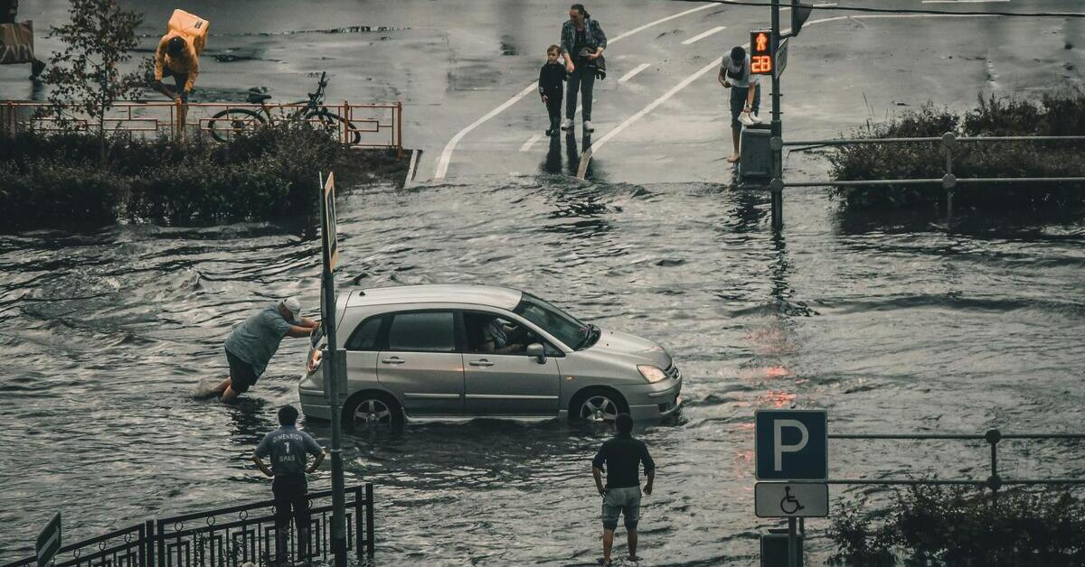 Überschwemmung Menschen stehen in Wasser, ein Mann schiebt ein Auto durch die überschwemmte Straße