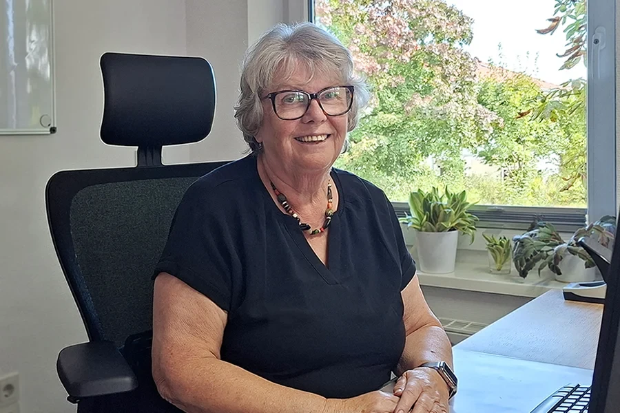 Petra Wandert sitzt im Büro vor einem Fenster mit Pflanzen im Hintergrund