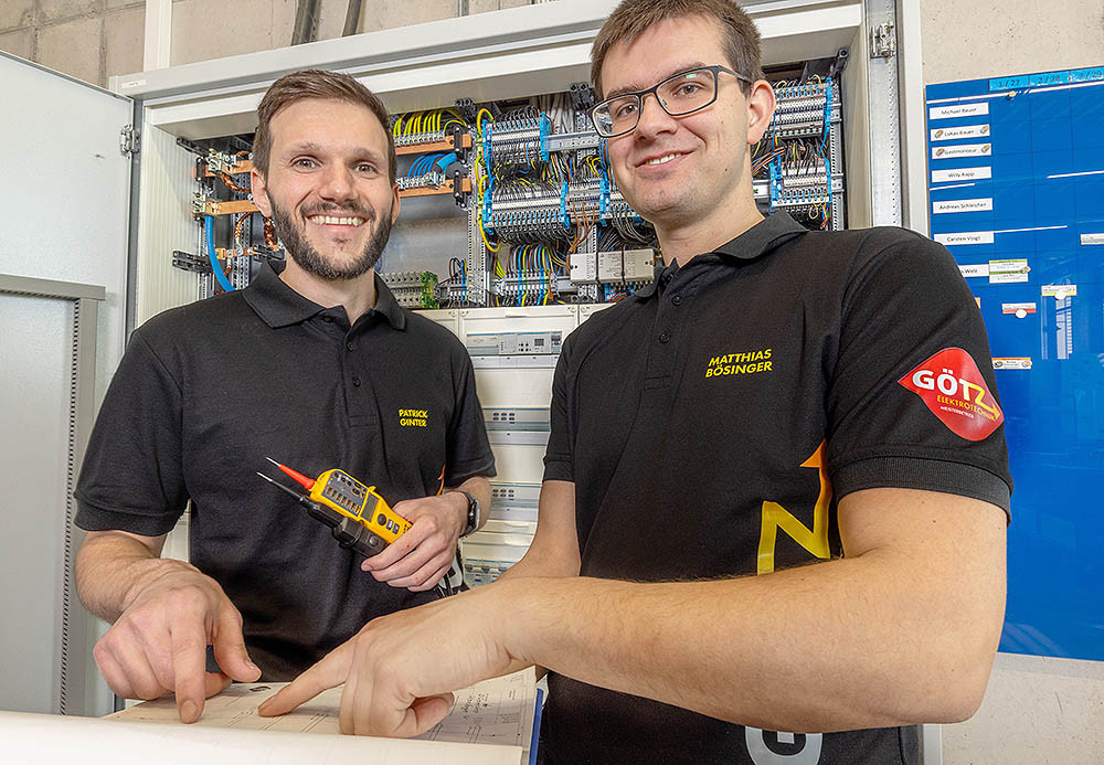 Zwei Elektriker, Patrick Gentner und Matthias Bösinger, prüfen einen Schaltschrank und besprechen technische Unterlagen.