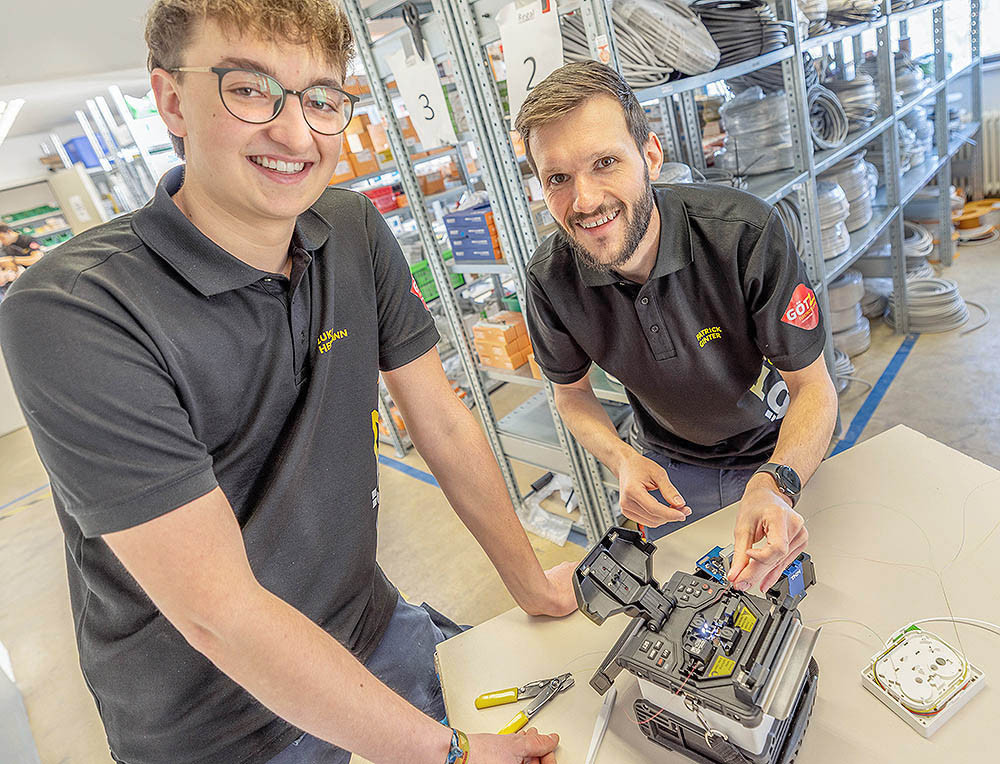 Zwei Techniker arbeiten gemeinsam an einem Gerät zur Kabelverbindung in einem Lager mit Regalen voller Elektromaterial.