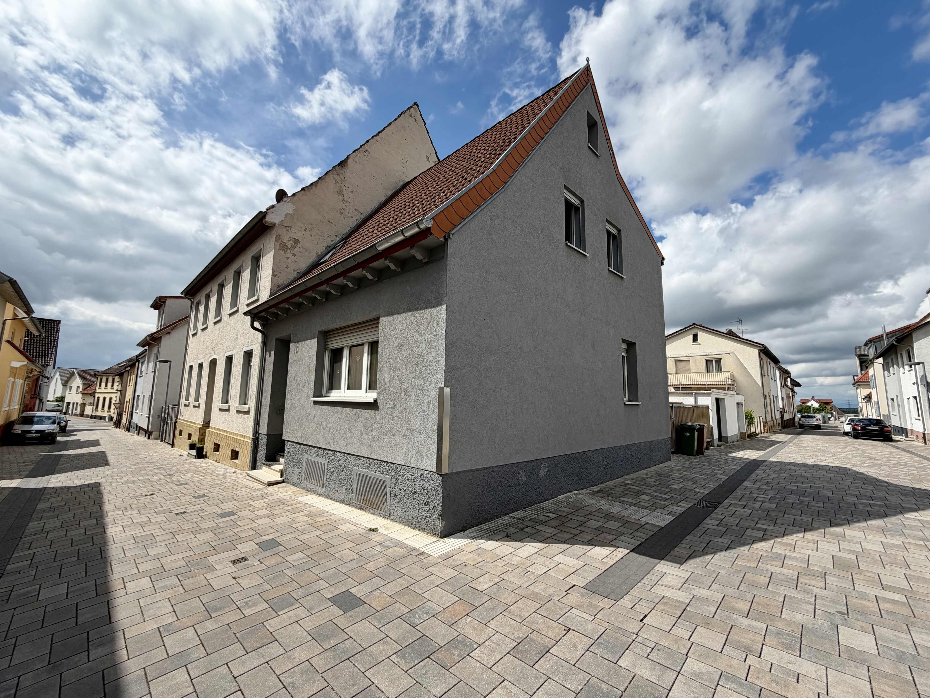 Wohnhaus an einer gepflasterten Straße mit grauer Fassade und Satteldach bei bewölktem Himmel
