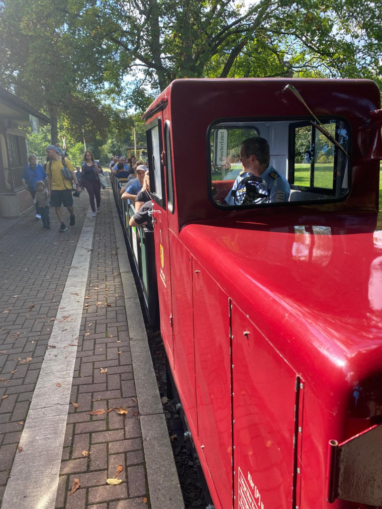 Rote Parkeisenbahn mit Fahrgästen an sonnigem Tag, Menschen laufen am Bahnsteig entlang