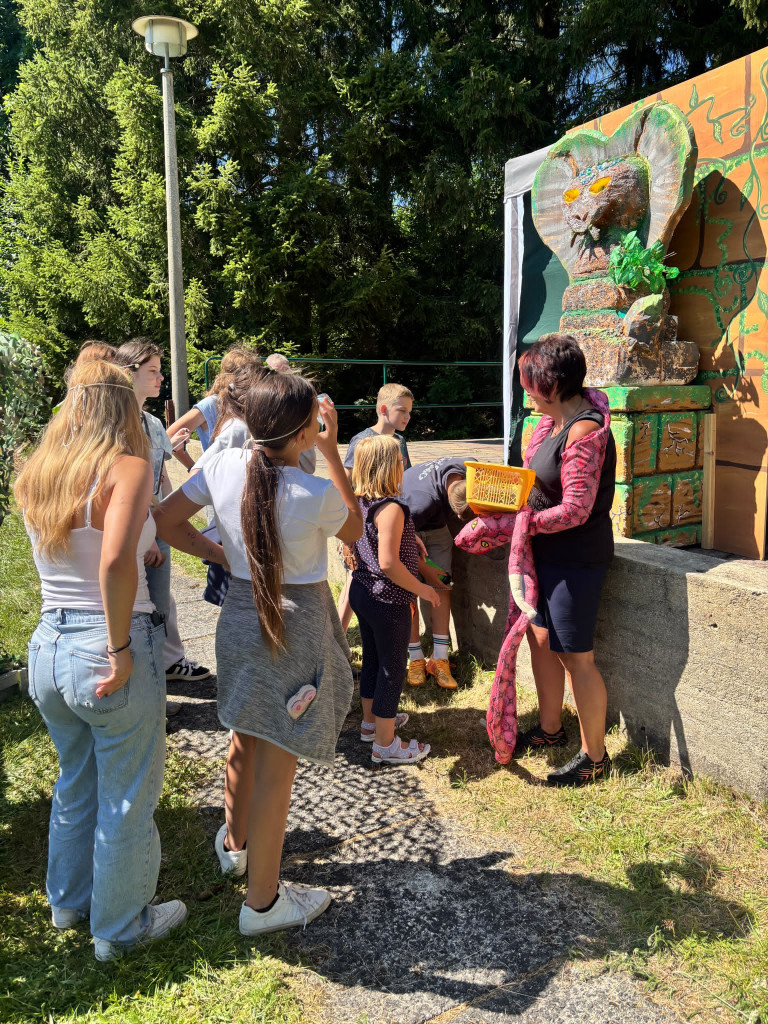 Kinder und Erwachsene erleben eine Outdoor-Aktion mit Schlangenfigur und Statue Kinder und Erwachsene bei einer Outdoor-Aktivität mit einer großen Schlangenfigur und einer dekorativen Statue im Hintergrund
