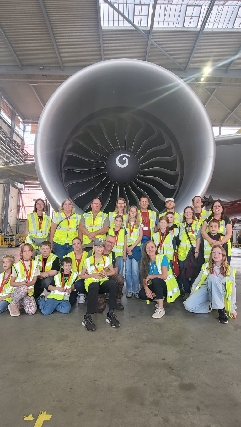 Gruppenfoto von Kindern und Erwachsenen in Warnwesten vor einem großen Flugzeugtriebwerk in einer Flugzeughalle