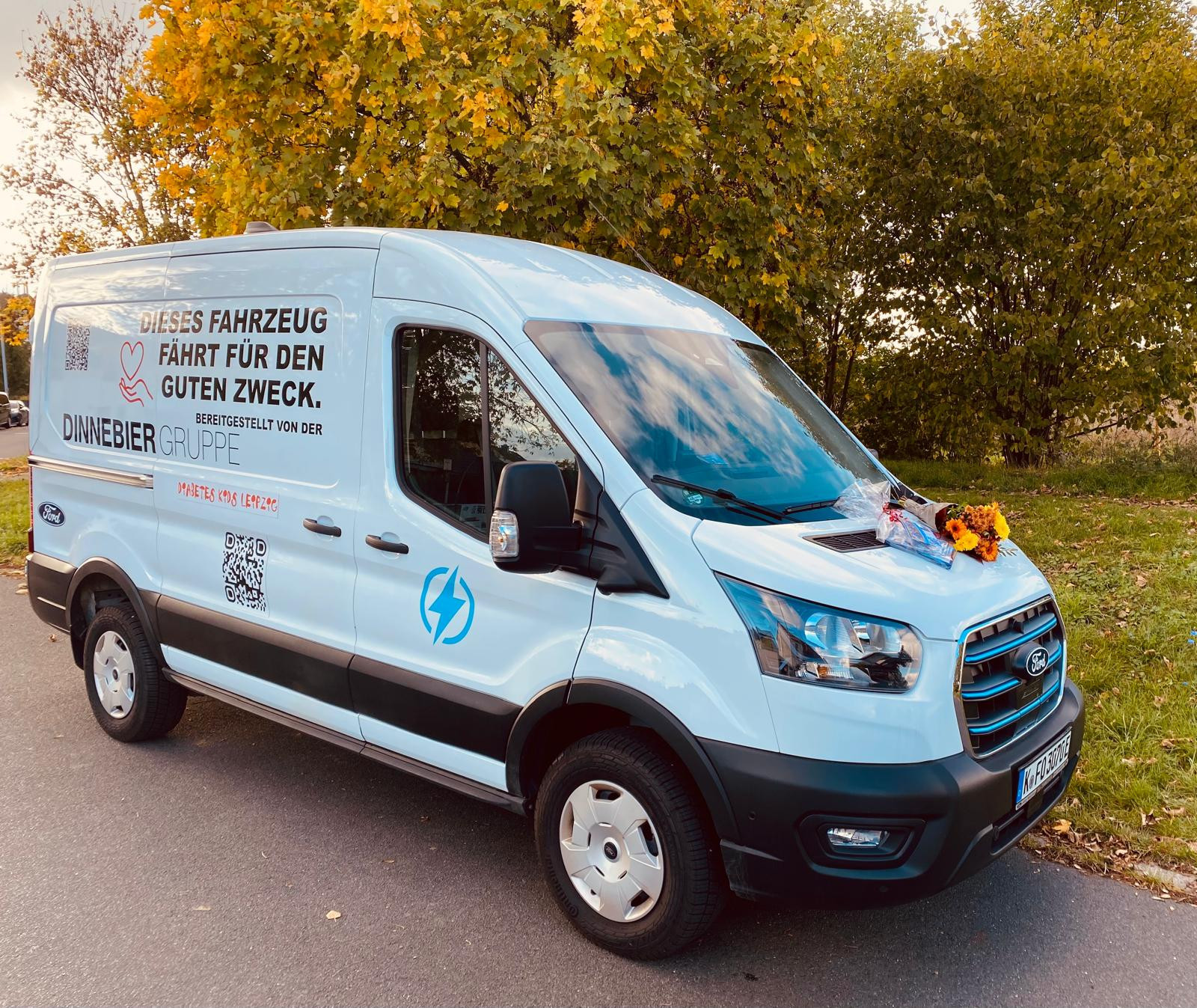 Elektro-Transporter mit Aufschrift 'Dieses Fahrzeug fährt für den guten Zweck' und Blumenstrauß auf der Motorhaube, bereitgestellt von der Kneibier Gruppe