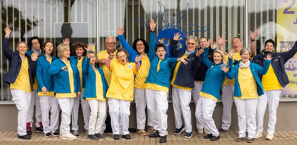 Teamfoto einer fröhlichen Gruppe von Mitarbeitenden in gelber und blauer Arbeitskleidung vor einem Firmengebäude