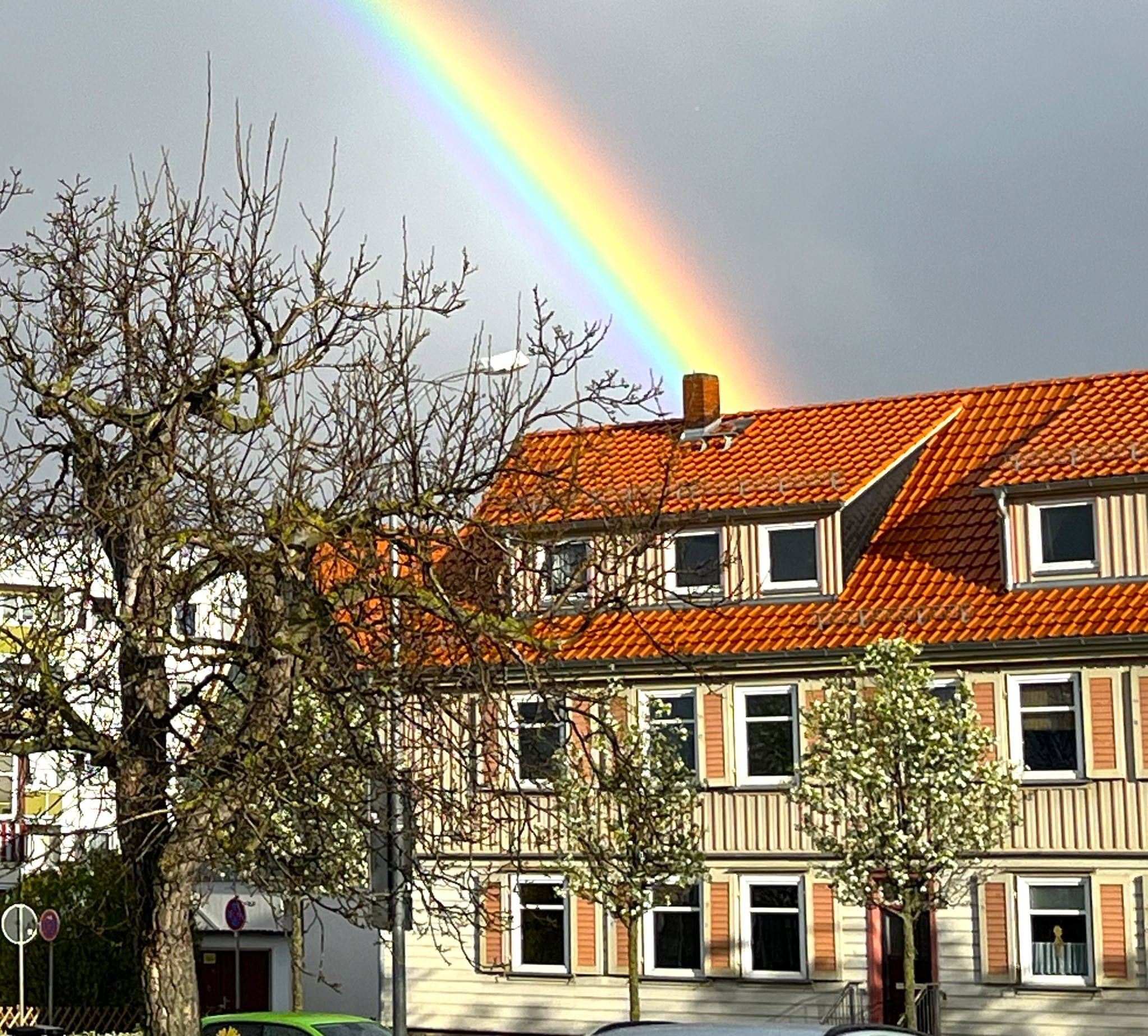 Regenbogen über einem Haus mit rotem Ziegeldach und blühenden Bäumen, Frühlingsstimmung
