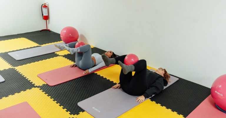 Zwei Frauen machen Fitnessübungen mit einem Gymnastikball auf Matten im Fitnessstudio.