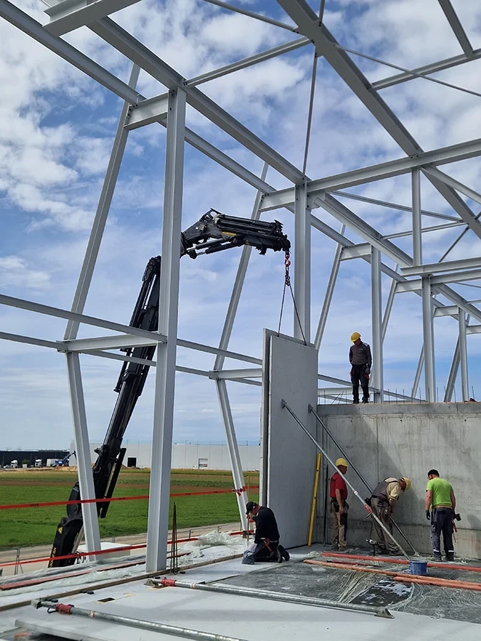 Krandienst hebt Betonelement bei Hallenbau – Bauarbeiter montieren Wand an Stahlkonstruktion