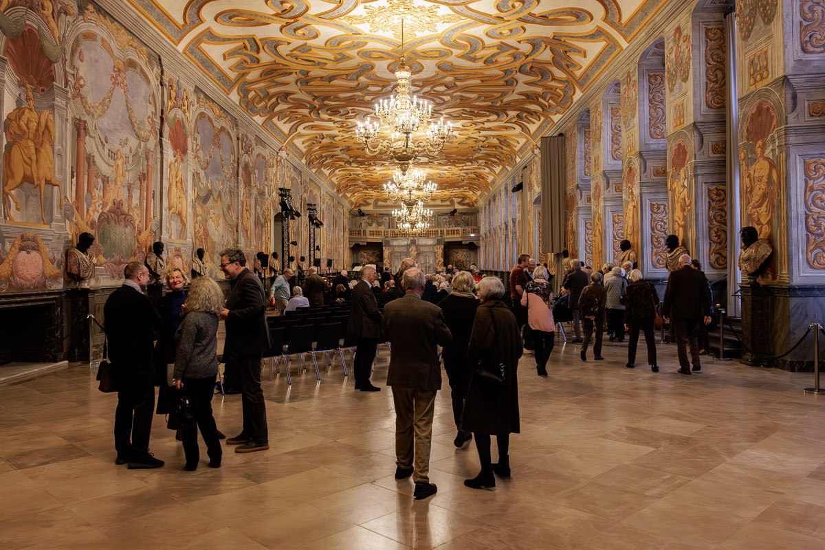 Menschen bei einer Veranstaltung in einer prunkvollen Galerie mit barocker Decke und Wandmalereien