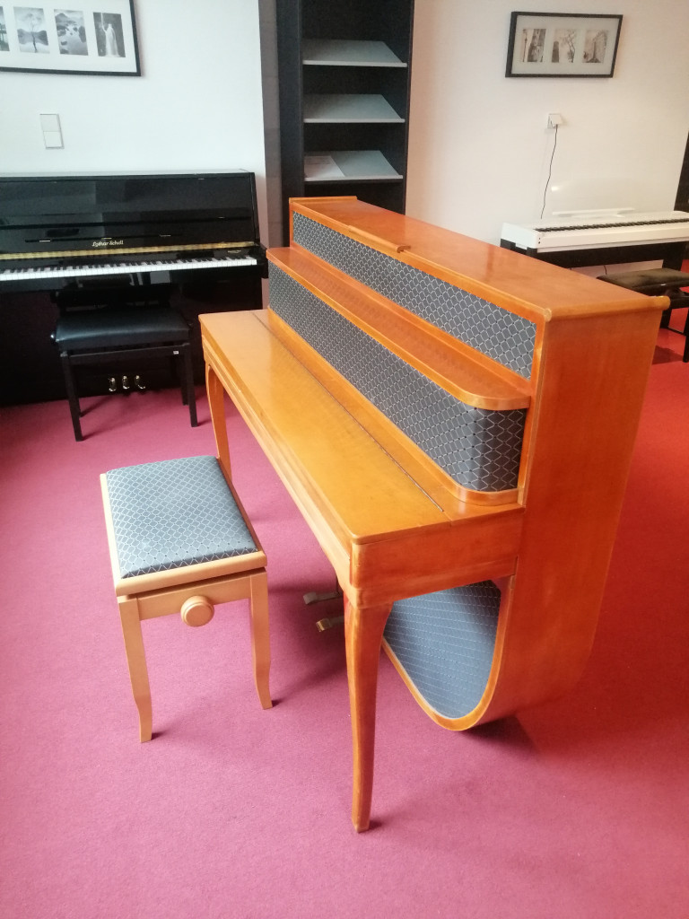 Helles Klavier mit passender Sitzbank in einem Musikraum mit rotem Teppichboden
