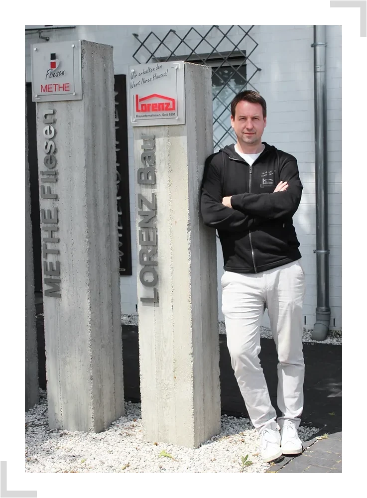 METHE Fliesen und LORENZ Bau Firmenschilder mit Person auf dem Firmengelände Person steht vor Betonsäulen mit den Aufschriften METHE Fliesen und LORENZ Bau, daneben Firmenschilder von Methe und Lorenz Bau
