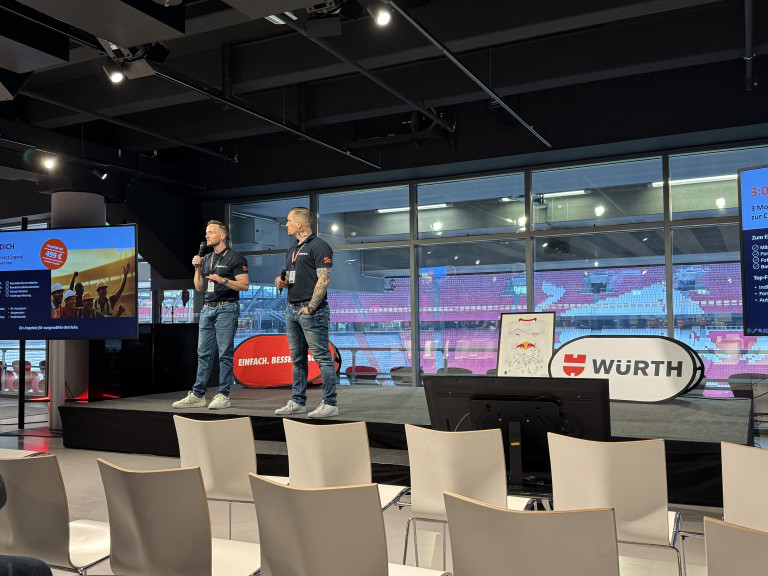 Zwei Moderatoren auf einer Bühne bei einer Würth-Veranstaltung im Stadion, mit Red Bull Leipzig Trikot im Hintergrund